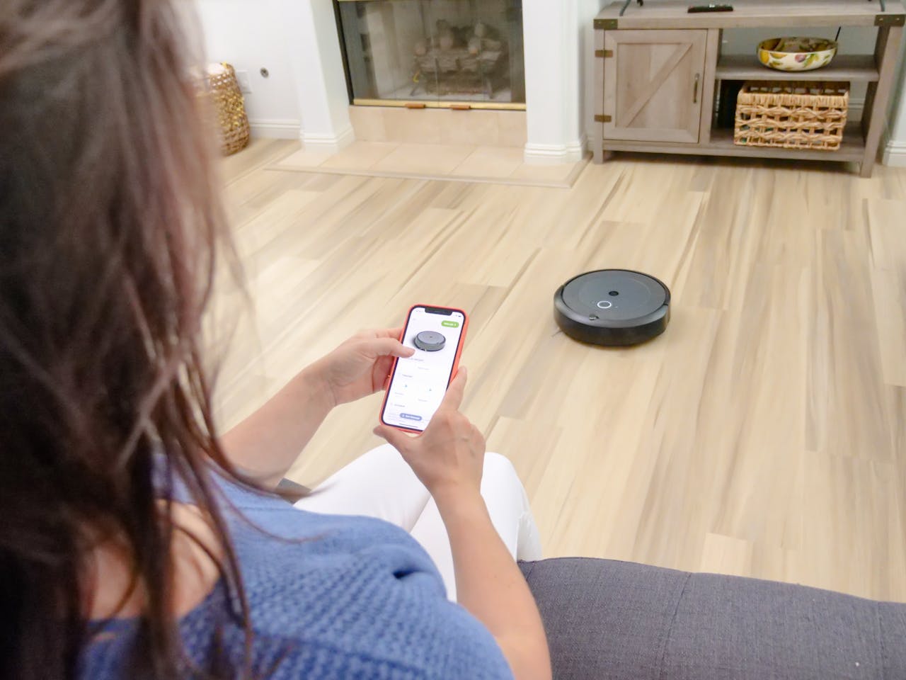 hero-02 Overhead view of a woman controlling a robot vacuum using a smartphone app on a wooden floor.